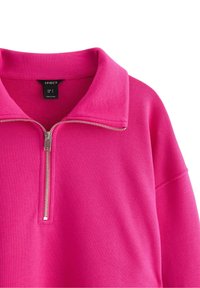 Lys pink pullover med krave og en sølvfarvet lynlås, der er delvist åbnet, så indersiden med mærket og syningsdetaljer vises.