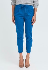 Blaue, verjüngte Hose mit einem Kordelzugbund, die Falten und eine glatte Textur aufweist, kombiniert mit nude-farbenen High Heels vor einem einfarbigen Hintergrund.