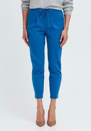 Blue tapered pants with a drawstring waist, featuring pleats and a smooth texture, paired with nude high heels on a plain background.