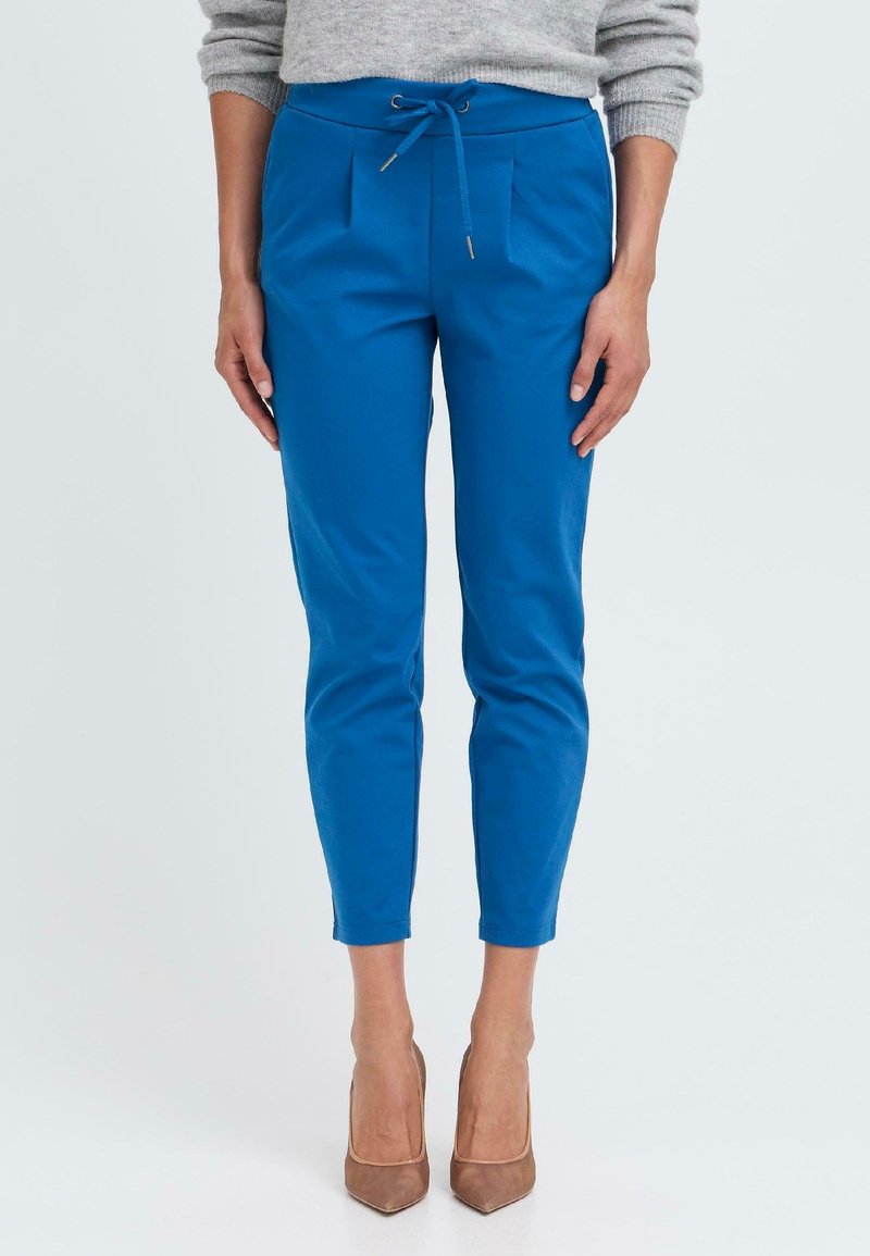 Blaue, verjüngte Hose mit einem Kordelzugbund, die Falten und eine glatte Textur aufweist, kombiniert mit nude-farbenen High Heels vor einem einfarbigen Hintergrund.