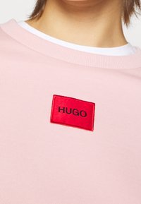 Sudadera rosa con cuello acanalado, que presenta una etiqueta roja rectangular con "HUGO" en texto negro. Textura simple y lisa.