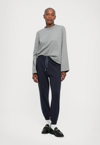 Person som står i en grå langermet skjorte, marineblå joggebukser med snor, hvite sokker og svarte loafers mot en ensfarget bakgrunn.