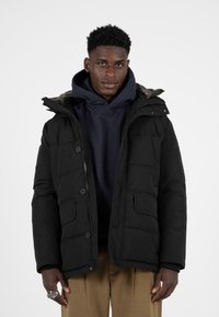 Černá prošívaná bunda s kapucí podšitou kožešinou, vybavená předním zipem, zapínáním na knoflíky a dvěma bočními kapsami. Nositá přes tmavě modrou mikinu.