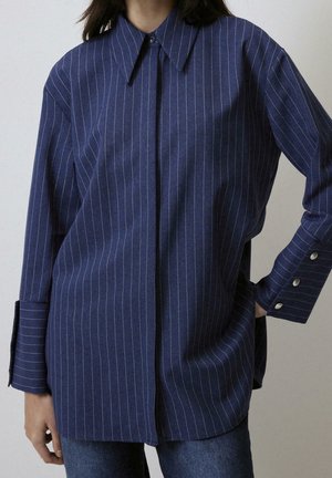 Personne portant une chemise bleue à fines rayures avec manches longues, col pointu et boutons de manchette argentés, associée à un jean bleu.