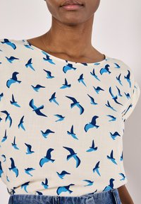Blouse à manches courtes en tissu crème clair avec un motif répété d'oiseaux bleus et marine, dotée d'un col rond et d'une texture douce.