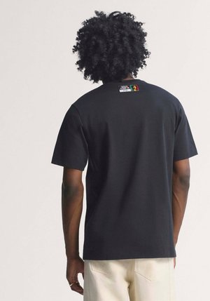 Mann med krøllete hår iført en svart kortermet skjorte med to små grafiske lapper nær halslinningen på baksiden.