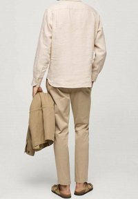 Mężczyzna ubrany w beżową koszulę z długim rękawem i spodnie, trzymający w jednej ręce jasnobrązową kurtkę, stojący boso w brązowych sandałach na jednolitym tle.