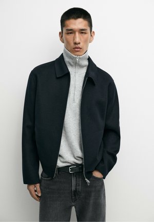 Homme portant une veste noire à fermeture éclair sur un pull gris clair à demi-fermeture éclair et un jean gris foncé, debout devant un fond blanc uni.