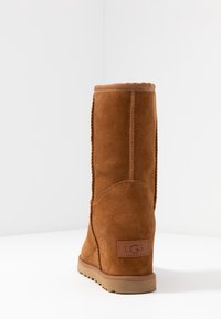 Botín UGG de ante marrón, con suela plana, visto desde la parte trasera sobre una superficie blanca y un fondo blanco liso.