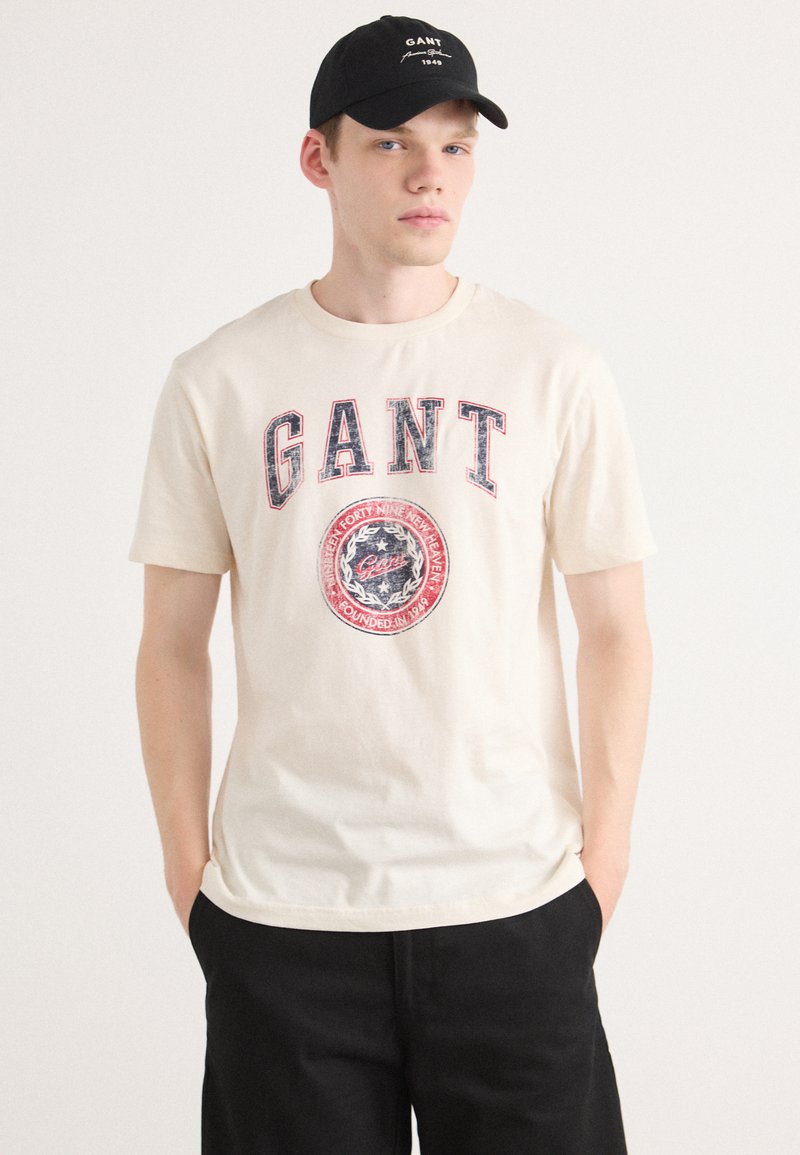 Cream-colored cotton T-shirt featuring a blue and red graphic with "GANT" text. Black cap with logo. Structured, casual fit.