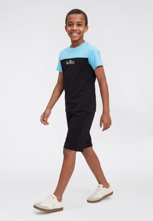 Ragazzo sorridente che cammina, indossa una maglietta a maniche corte azzurro chiaro e nera, pantaloncini neri e scarpe da ginnastica bianche su uno sfondo neutro.