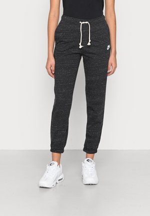 Person iført sorte melerede joggingbukser med hvid snor og hvide Nike Air Max sneakers, stående mod en ensfarvet baggrund.