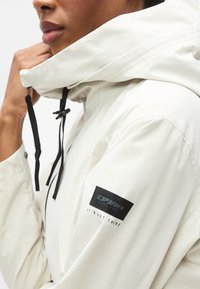 Λευκό αδιάβροχο μπουφάν με ρυθμιζόμενα μαύρα κορδόνια, ψηλό γιακά και μαύρο λογότυπο στο μανίκι. Λείες επιφάνειες χωρίς υφή.