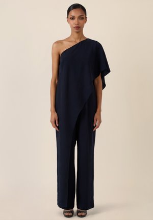 Frau trägt einen marineblauen Ein-Schulter-Jumpsuit mit weitem Bein und schwarzen offenen Sandalen, steht vor einem schlichten beigen Hintergrund.