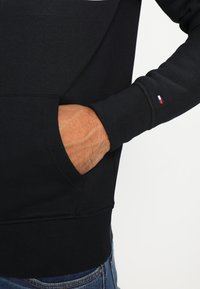 Felpa nera con una texture morbida, polsini a costine e tasca frontale. Presenta un piccolo logo colorato sulla manica.