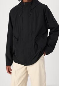 Czarna wodoodporna kurtka z kapturem, wyposażona w zamek błyskawiczny z przodu, zapięcie na zatrzaski oraz boczne kieszenie. Gładka faktura i luźny krój.