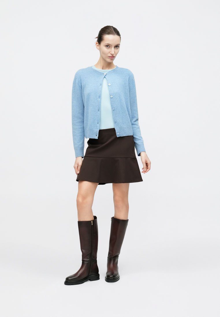 Cardigan tricoté bleu clair, encolure ronde, boutons sur le devant ; associé à un haut bleu clair et une jupe évasée marron. Le mannequin porte des bottes marron montant jusqu'aux genoux.