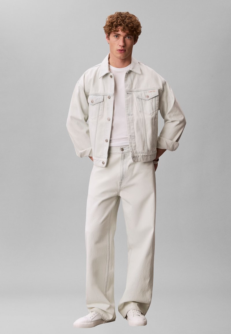 Νεαρός με σγουρά μαλλιά που φοράει ανοιχτόχρωμο τζιν μπουφάν και παντελόνι πάνω από λευκό T-shirt και λευκά αθλητικά παπούτσια, στέκεται με τα χέρια στις τσέπες.