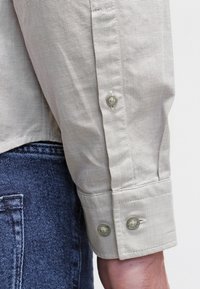Light gray striped shirt fabric with buttoned cuffs, featuring two green buttons and a smooth texture, paired with dark denim jeans.