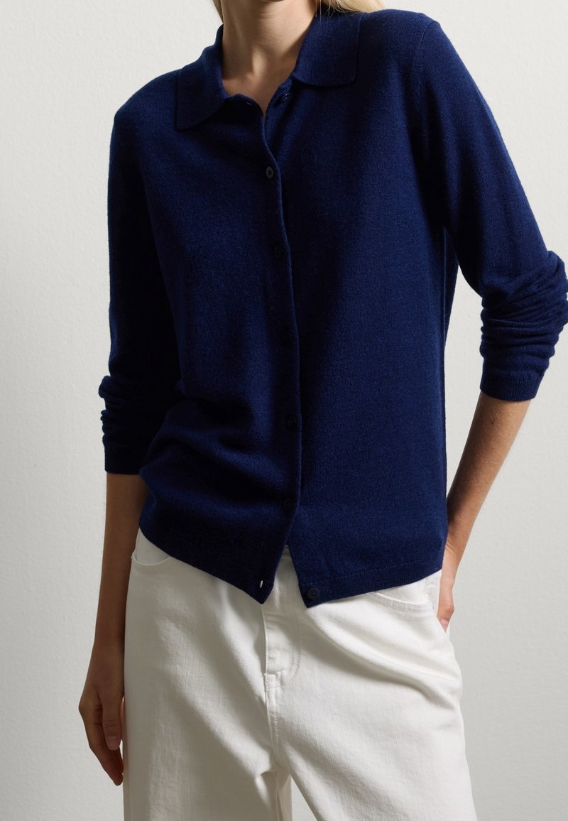 Femme portant un cardigan bleu marine boutonné avec un col et un pantalon blanc taille haute, debout avec une main dans la poche.
