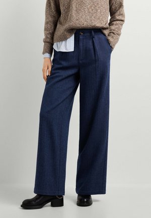 Person wearing wide-leg navy blue pants, beige sweater, striped shirt, and black heeled boots standing against a plain light background.