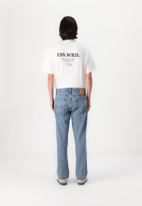 Tricou alb cu text negru, blugi bleu deschis și adidași gri. Tricoul are o grafică pe spate. Fundal simplu.