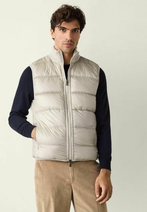 Mann trägt eine hellbeige gesteppte Weste über einem schwarzen Langarmshirt und braunen Cordhosen, steht vor einfachem Hintergrund.