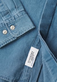 Poignet de chemise en denim bleu avec des boutons blancs et une étiquette blanche de la marque Superdry indiquant l'adresse et des numéros manuscrits.