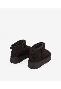 Botines de ante marrón con un suave forro de piel, punteras redondas, caña baja y suelas de goma. Incluyen tiradores traseros y una suela texturizada.