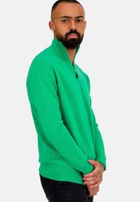 Grüner Strickpullover mit halb-zip Kragen, geripptem Saum und Ärmelabschlüssen, mit weichem Material und lockerer Passform, kombiniert mit dunklen Hosen.