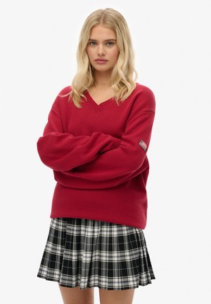 Jonge vrouw met blond haar, gekleed in een rode V-hals trui en een zwart-wit geruite rok, met gekruiste armen, op een neutrale achtergrond.