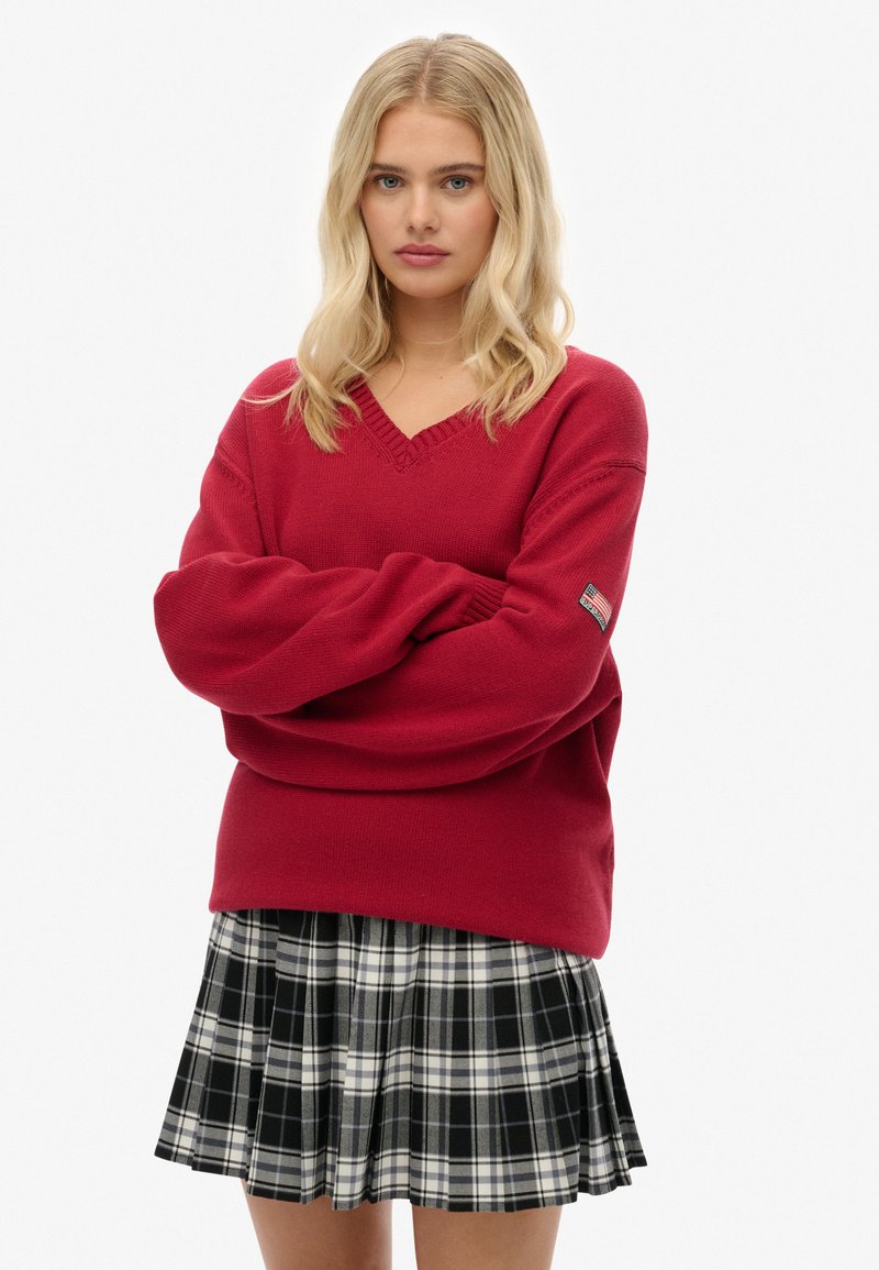Giovane donna con i capelli biondi, che indossa un maglione rosso a V e una gonna a quadretti bianco e nero, con le braccia incrociate, su uno sfondo neutro.