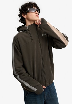 Ung mand iført mørk lynlåshættetrøje med lyse striber, solbriller og blå jeans, der kigger opad med hånden nær hagen.