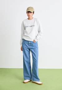 Grijze sweatshirt met "carhartt." in het blauw geprint, gecombineerd met lichtblauwe wijde jeans en een beige pet. Witte sneakers met bruine zolen.
