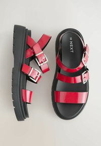 Sandales en cuir verni rouge avec plusieurs sangles réglables et boucles en argent. Semelle épaisse noire avec un motif de traction texturé.