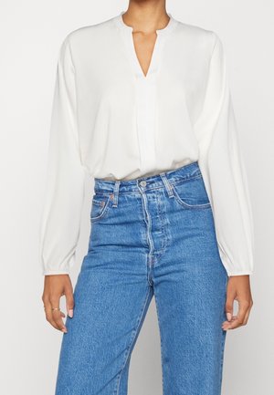Person wearing a white long-sleeve blouse with a V-neckline tucked into high-waisted blue denim jeans.