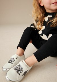 Vit läderskor med svarta leopardmönstrade detaljer och silverfärgade tåskydd. Bärs av ett barn som sitter, i kombination med svarta ribbade leggings.