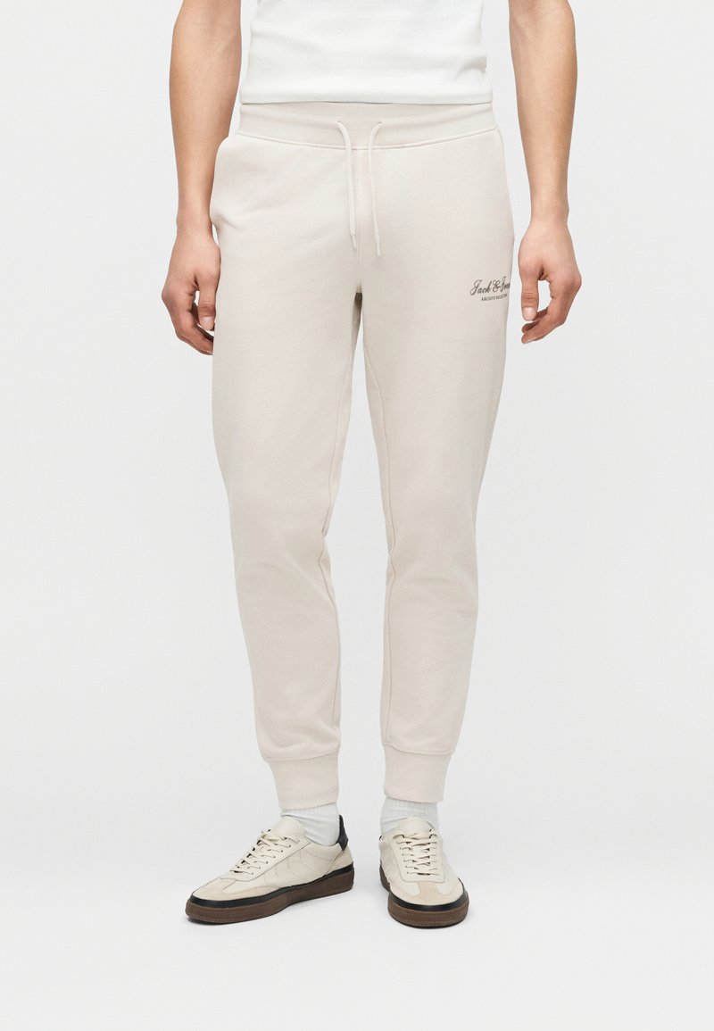 Άνδρας που φοράει μπεζ φόρμες με κορδόνι στη μέση και μανσέτες στους αστραγάλους, συνδυασμένες με λευκά αθλητικά παπούτσια και κάλτσες σε απλό φόντο.