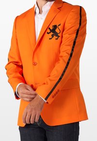 Pomarańczowy żakiet z czarnym haftem lwa na klatce piersiowej, kontrastowe czarne paski wzdłuż rękawów oraz gładka faktura materiału.