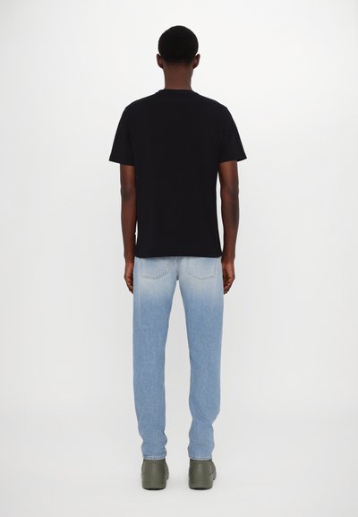 T-shirt en coton noir avec un col rond, associé à un jean léger bleu délavé et des chaussures vertes. Le cadre est simple et neutre.