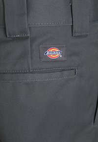 Szare spodnie robocze wykonane z trwałego materiału, z tylną kieszenią i naszywką z logo Dickies w kolorach czerwonym, żółtym i niebieskim. Widziane detale przeszyć.