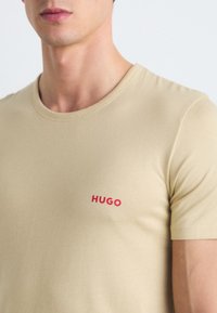Beige bomull T-skjorte med rund hals, tettsittende fasong, og en liten rød "HUGO"-logo på venstre bryst. Glatt tekstur med korte ermer.