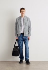 Jachetă bomber gri deschis peste un tricou alb, blugi și pantofi negri; ținând o geantă neagră; stând în fața unui fundal alb.