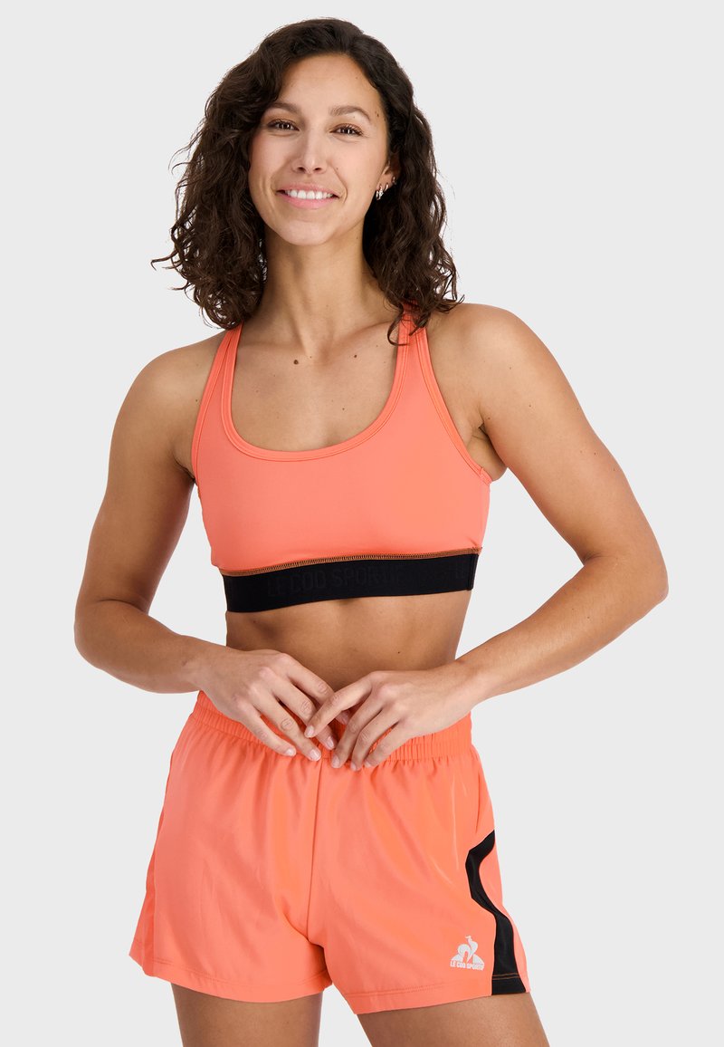 Športový outfit pozostávajúci z jasne koralového športového podprsenky s čiernym pásom a zodpovedajúcich koralových šortiek s čiernym bočným pruhom a logom.