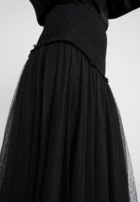 Falda negra con una cinturilla fruncida texturizada, en capas con tul semitransparente adornado con patrones sutiles y un dobladillo con volantes.