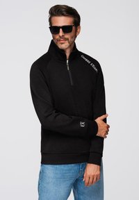 Sudadera negra con cremallera y alto cuello, textura suave, detalles de logo en blanco y corte holgado, combinada con unos jeans de color azul claro.