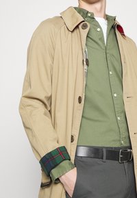 Tan trenchcoat över en grön skjorta, med en rutigt accent på ärmen. Outfiten inkluderar ett svart bälte och grå byxor.