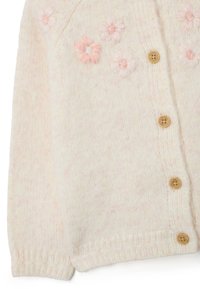 Gilet en tricot crème avec broderie florale rose, boutons en bois naturel et poignets côtelés pour une texture supplémentaire.