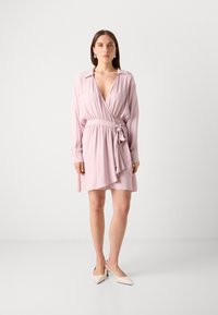 Light pink wrap dress with long sleeves, deep V-neck, elastic waist, and front tie detail. Smooth fabric with a flowing skirt.
