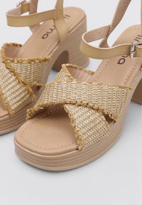 Tan platform sandals with woven straw straps, featuring frayed edges, an ankle strap, and a textured sole. Smooth beige footbed.
